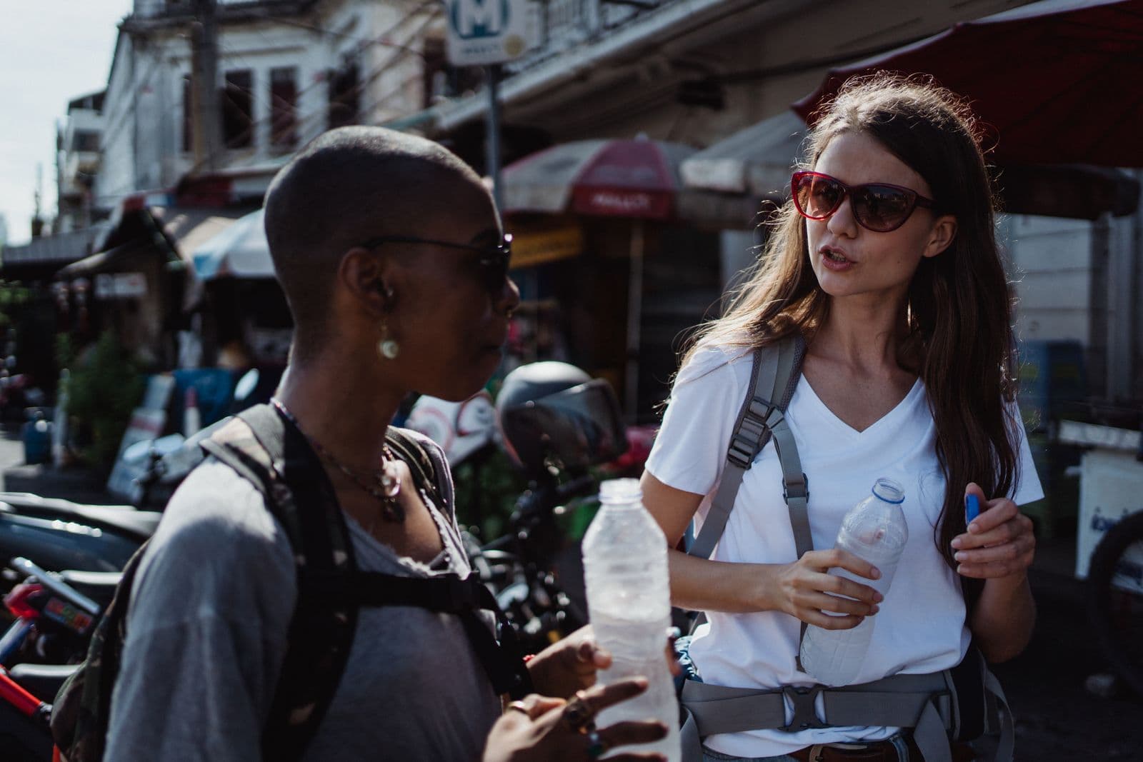 Two friends holding bottled drinks and talking at a street market