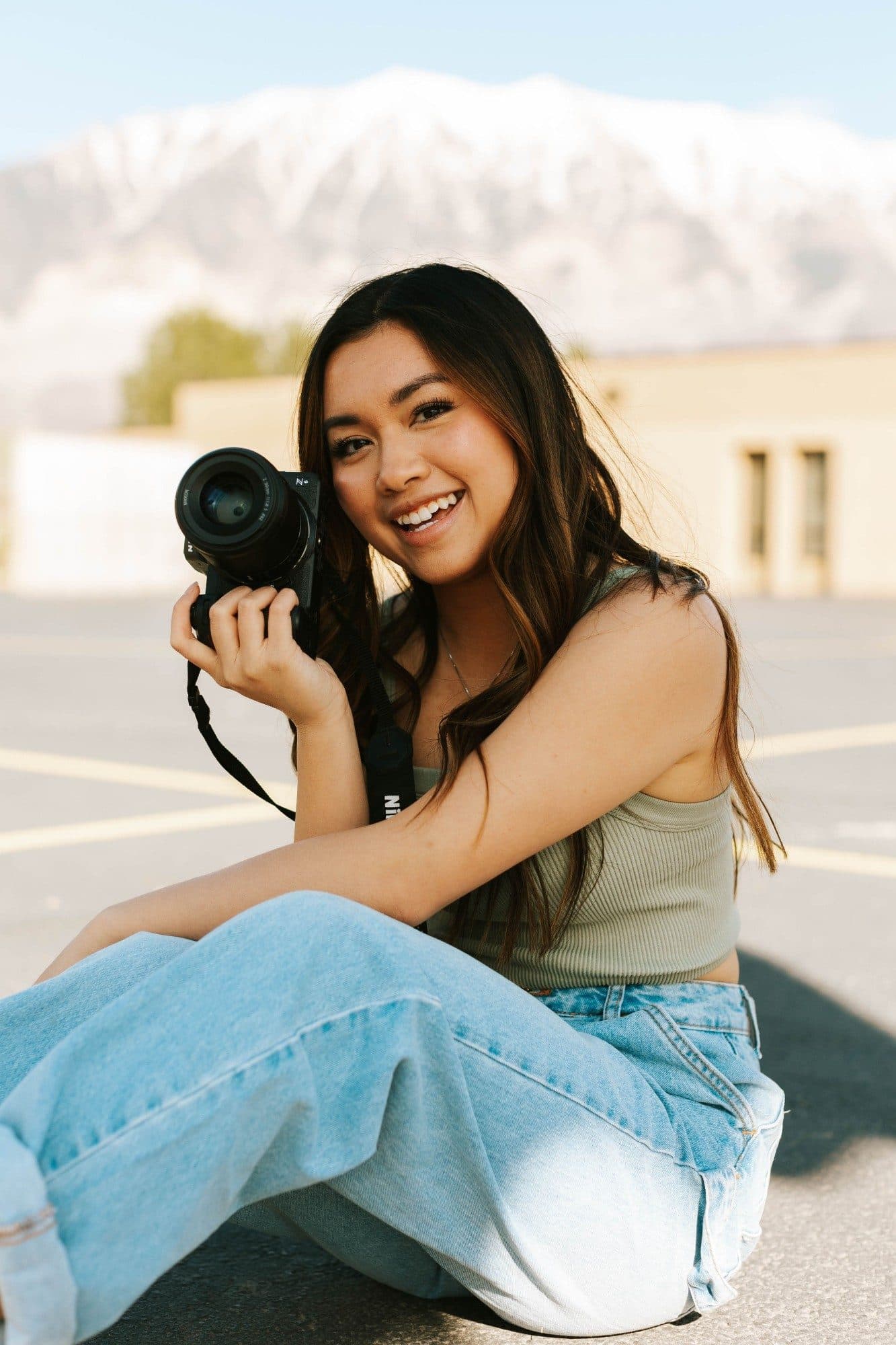 Angel Phommachan sitting on pavement with a Nikon camera, laughing, snowy mountains behind her