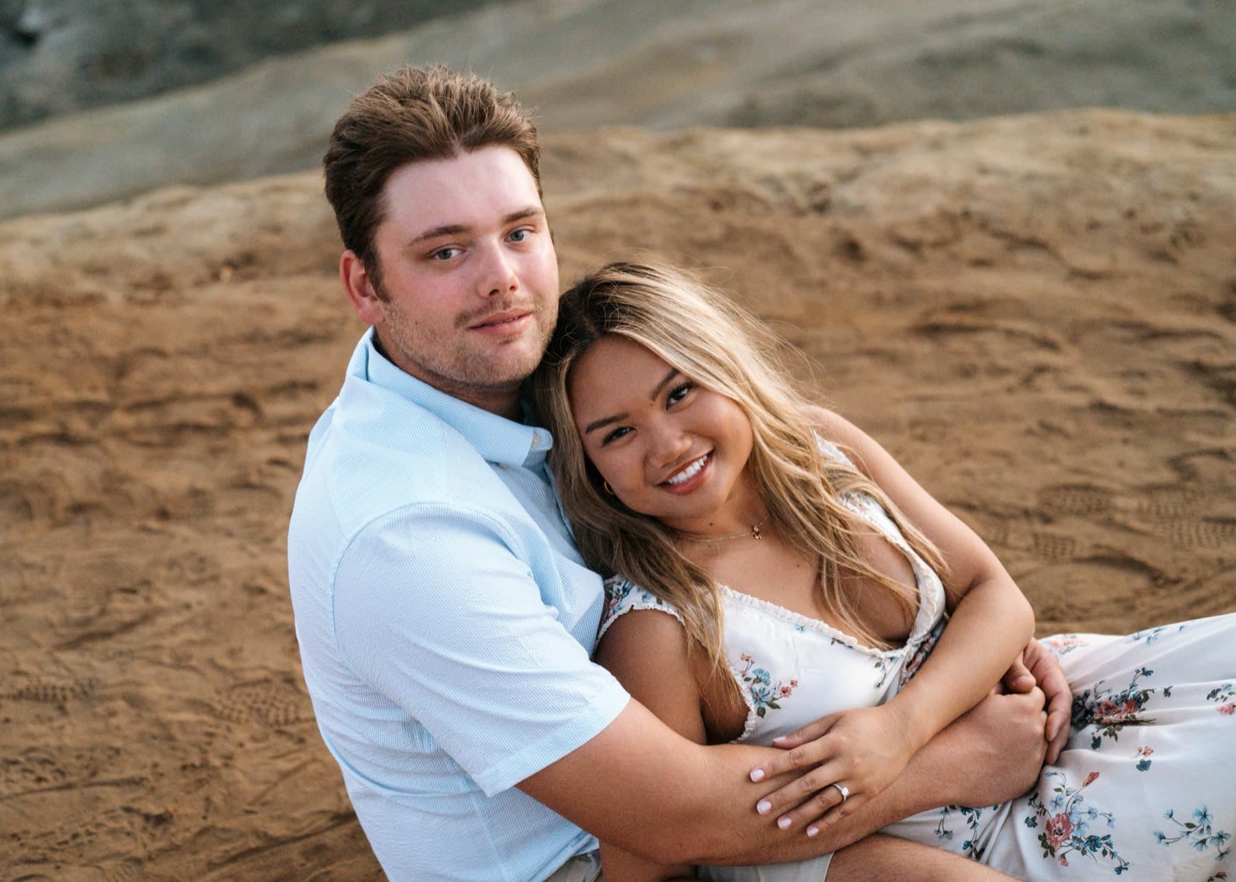 Engaged couple sitting on the sand by the ocean, laughing as he kisses her cheek