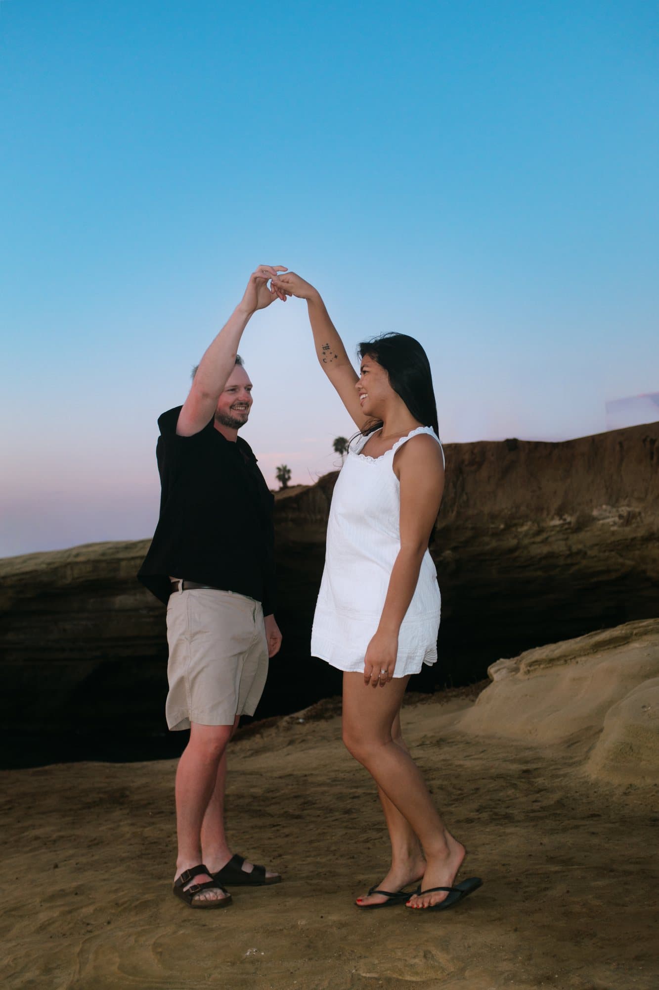 Couple on a cliff edge with their hands raised together against a blue sky