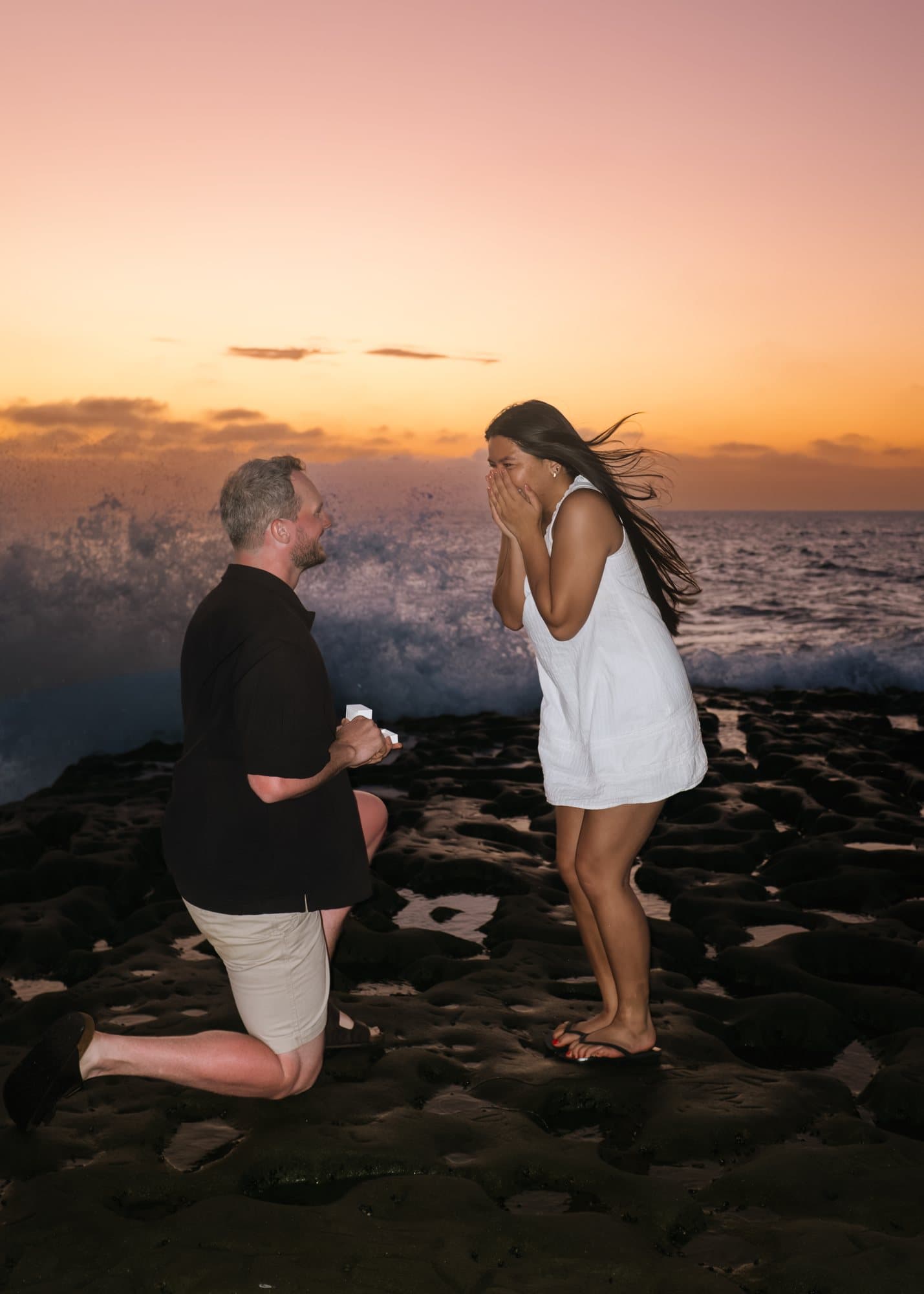 Proposal on the rocks at sunset as a wave breaks behind the couple