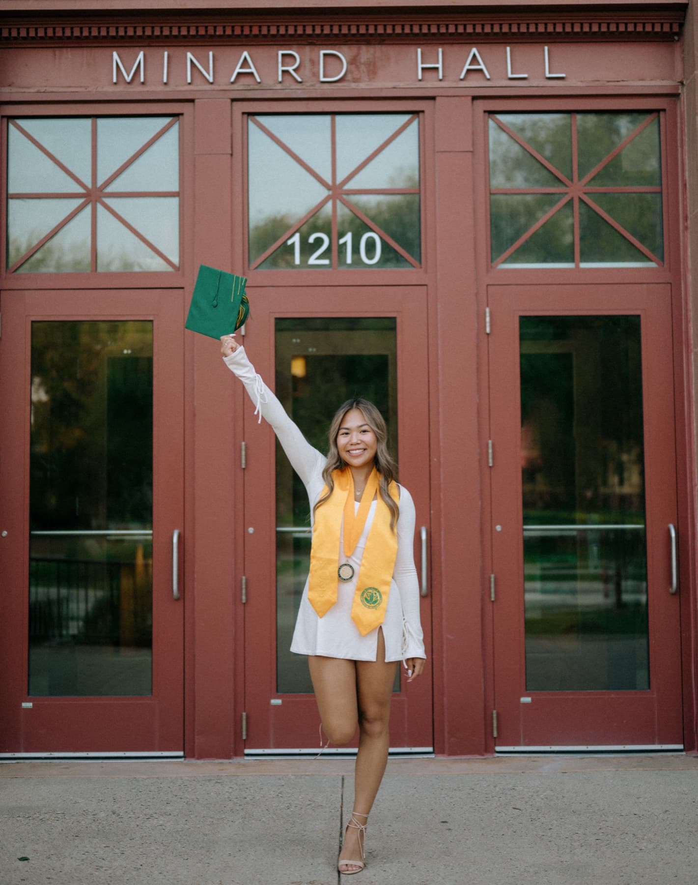 Graduate in a gold stole lifting her cap outside Minard Hall